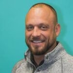 A man with a trimmed beard and short hair smiles confidently at the camera, wearing a light gray zip-up sweater against a solid teal background.