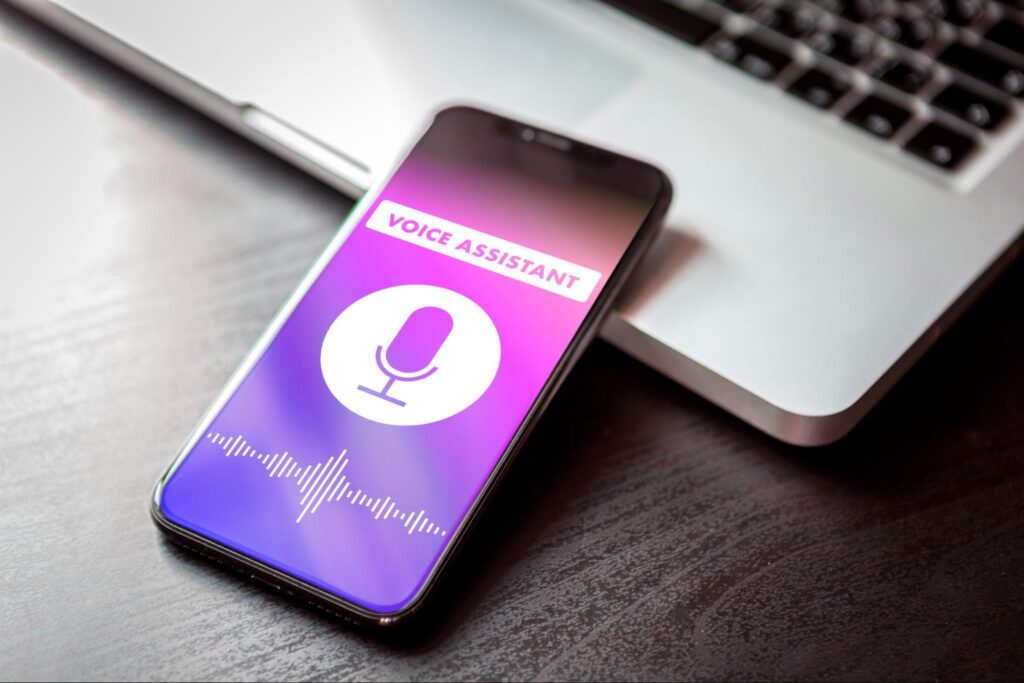 Smartphone with AI Voice Search assistant open, featuring microphone and sound waves, on dark desk beside laptop keyboard.