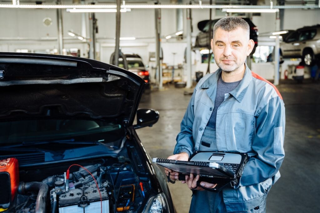 A mechanic in a gray uniform stands beside an open-hood car, using a diagnostic tool—demonstrating the professional approach found in top electrician jobs. The auto repair shop is equipped with multiple vehicles and tools, reinforcing Hook Agency’s focus on industry expertise and readiness.