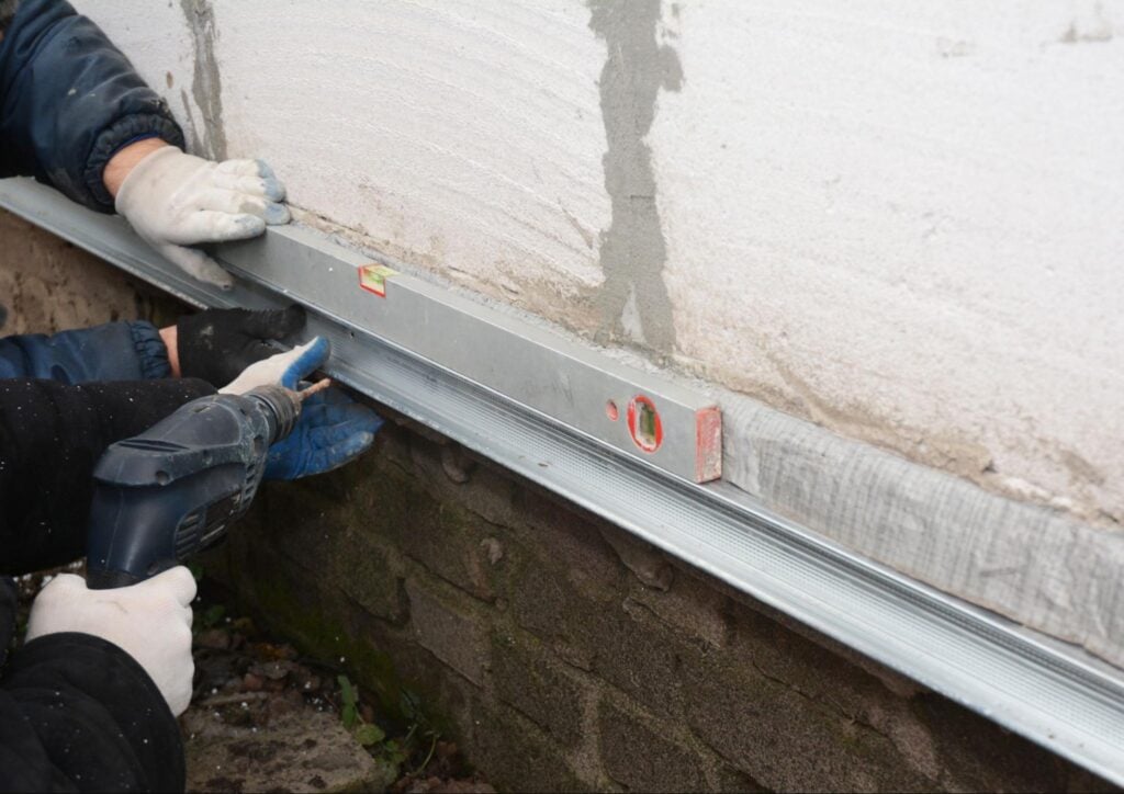 Gloved professionals use a spirit level and drill to precisely secure a metal strip at an exterior wall base, ensuring accuracy.