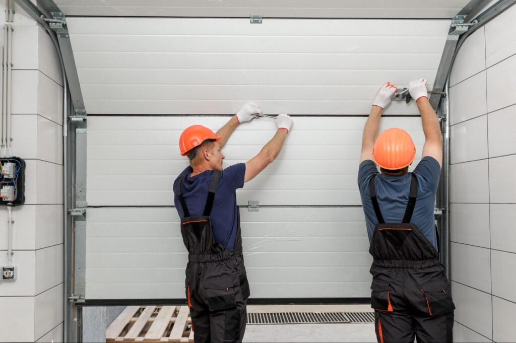 Two workers in orange helmets and dark overalls install a white garage door, using tools inside a garage for Hook Agency clients.