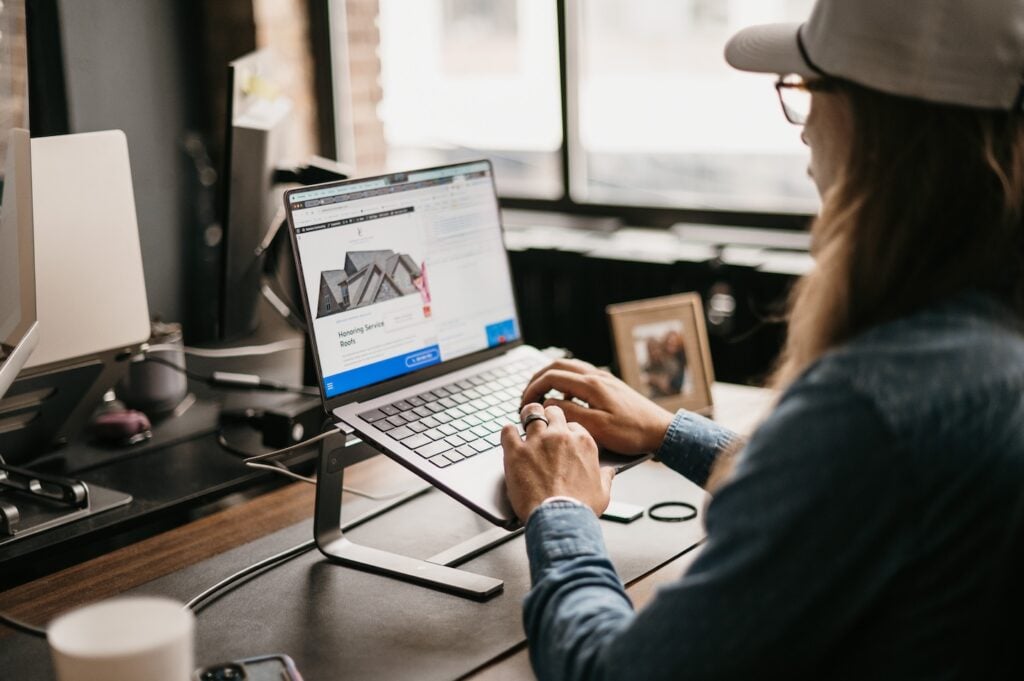 Professional works intently on content quality at a laptop, surrounded by a monitor, photo frame, coffee cup, and window.