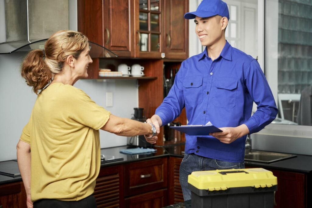 A woman in a yellow shirt confidently shakes hands with a technician in a blue uniform and cap, who holds a clipboard. They stand amid dark wooden kitchen cabinets, discussing effective solutions to an issue. A yellow toolbox on the counter signifies that no job is too complex for the trusted experts in our referral program.