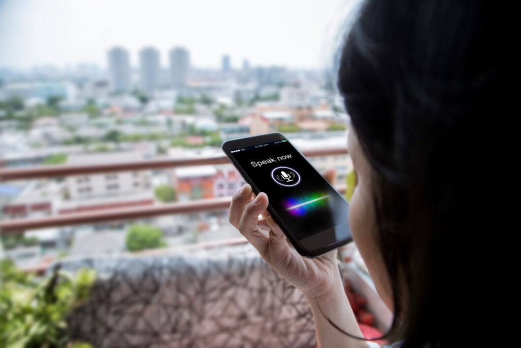 A person stands on a balcony, smartphone in hand displaying a voice search app's "Speak now" prompt. The cityscape of towering buildings stretches beyond, symbolizing how home services businesses can leverage technology to enhance customer connections.