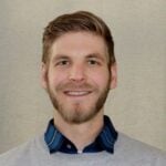 A man with short brown hair and a beard smiles warmly at the camera, embodying the soothing charm of Aquarius Home Services. Dressed in a gray sweater over a blue collared shirt, he exudes confidence against a neutral background.