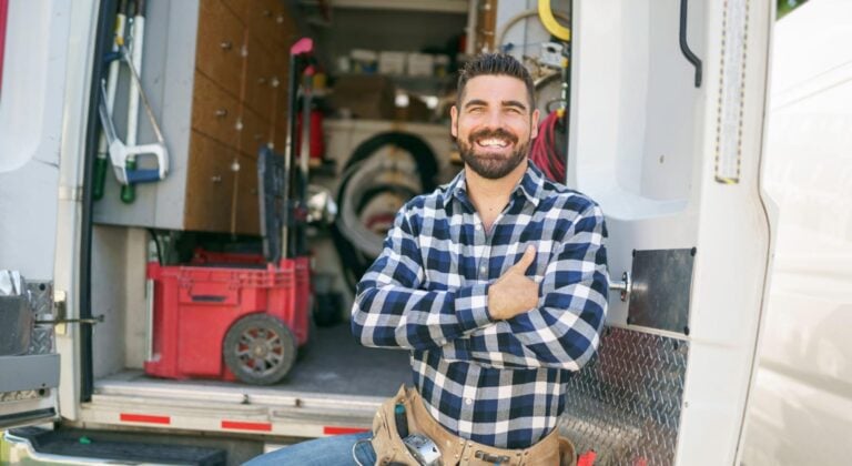 Professional electrician in plaid shirt and tool belt stands confidently at work van door, perfect for Hook Agency marketing.
