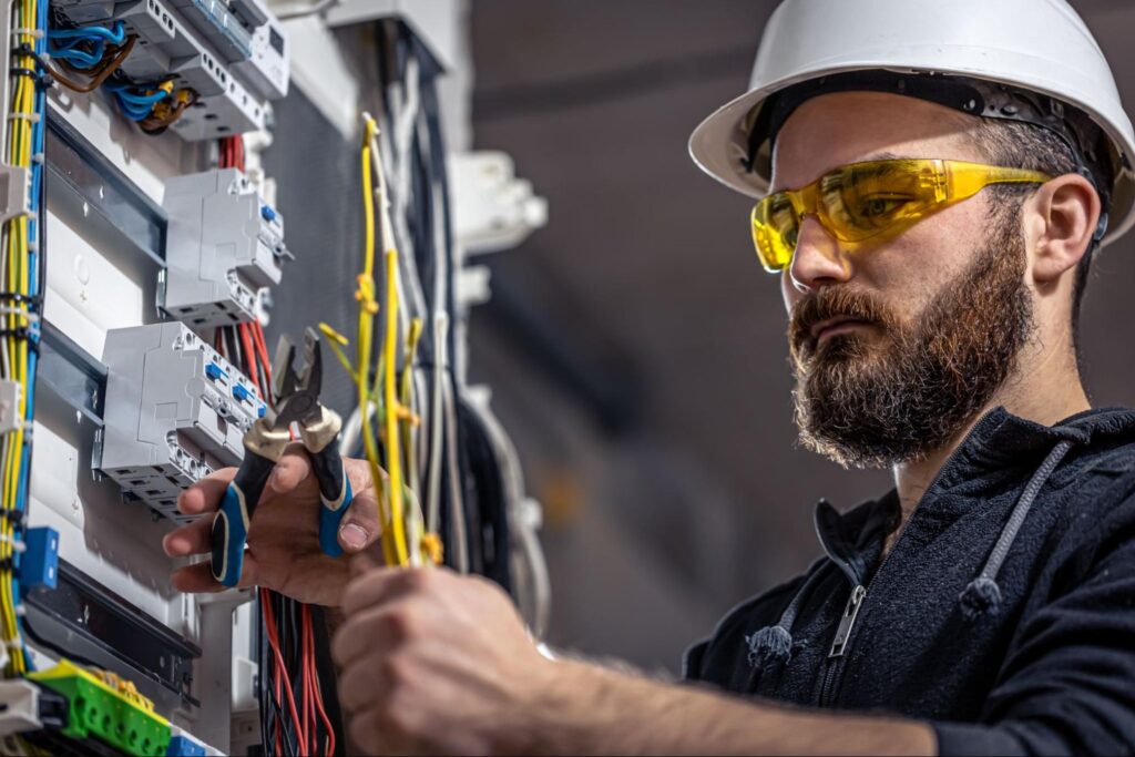 Experienced electrician in white hard hat and yellow safety glasses uses pliers on a control panel, providing expert home services.
