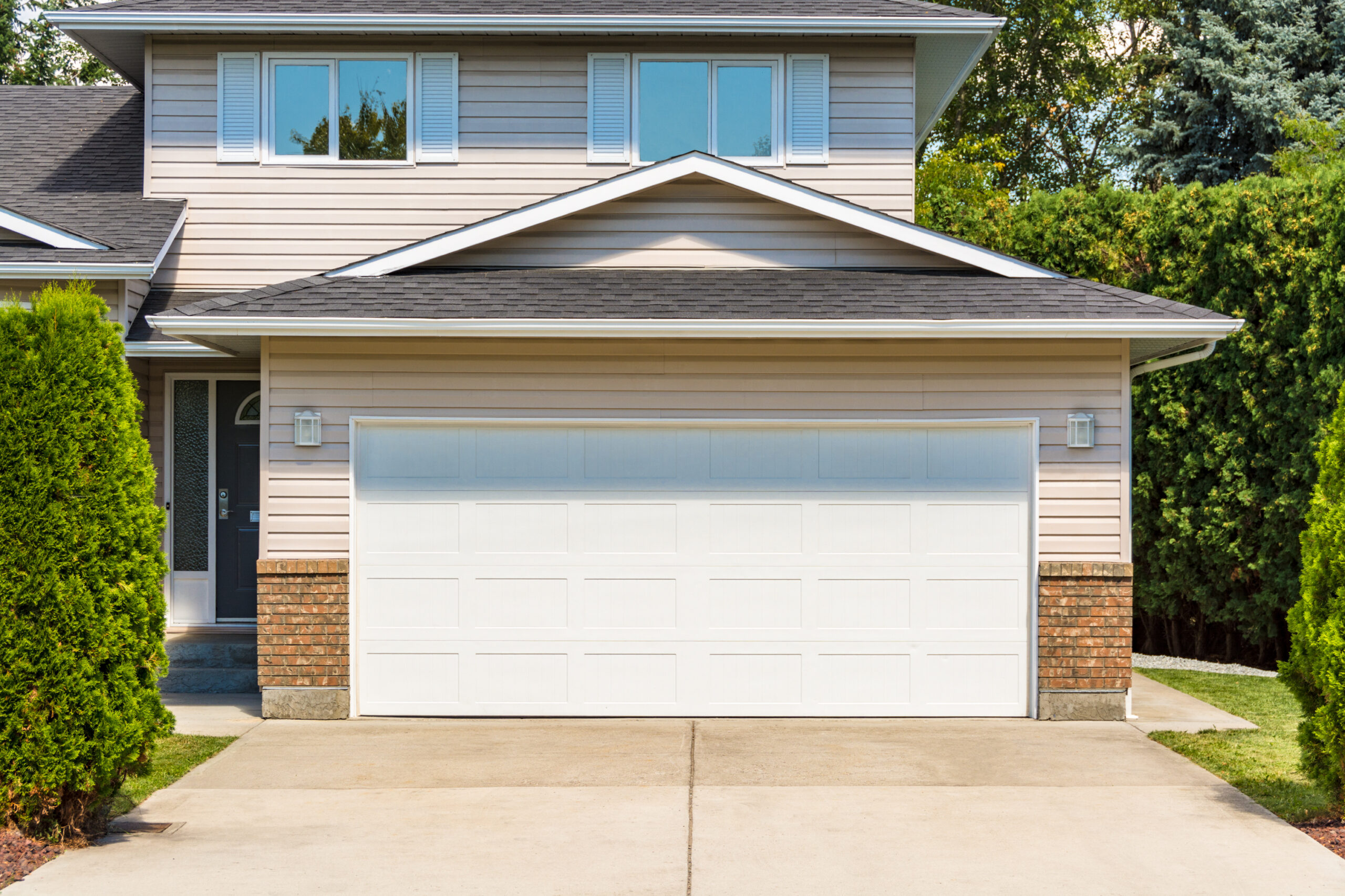 Enhance your home's curb appeal with our sleek garage floor coating. This two-story house, featuring beige siding and a prominent white garage door, is complemented by a tidy concrete driveway lined with lush green bushes. Upper-floor windows and a sophisticated gray roof complete this modern design.