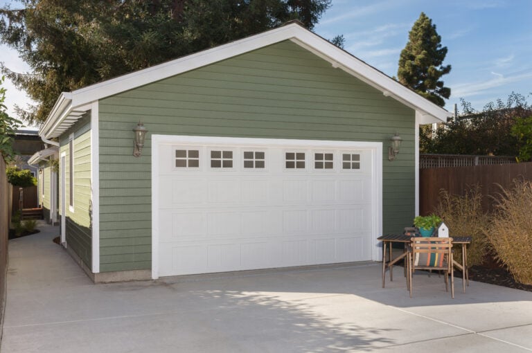 A green detached garage features a clean white sectional door and two wall lights, offering an impressive focal point. Adjacent is a charming outdoor seating area with a small table, two chairs, and a potted plant, all set against the backdrop of tall trees and a clear blue sky. Ideal for construction enthusiasts who appreciate craftsmanship in their surroundings.