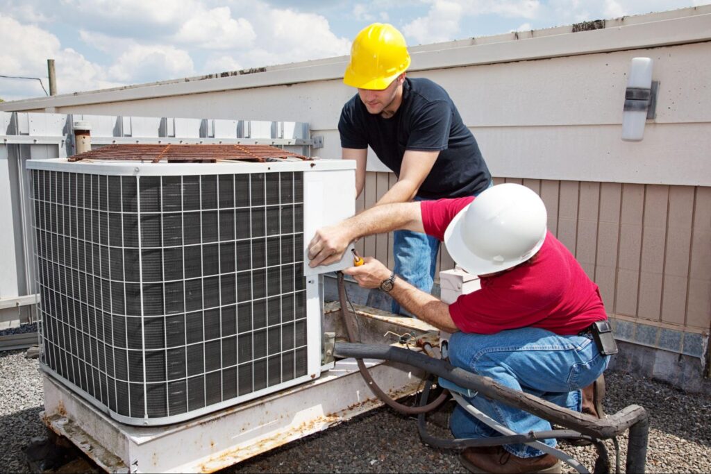 Two trades workers in hard hats repair a rooftop AC unit, using tools and teamwork for essential year-round maintenance.