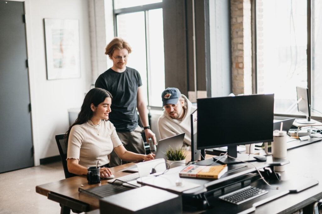 In a modern office, three people gather around a desk. A woman sits smiling at a laptop while two men stand nearby, discussing strategies for the best HVAC marketing agency of 2024. The setup includes monitors, a plant, and office items. Large windows bathe the art-adorned walls in natural light.