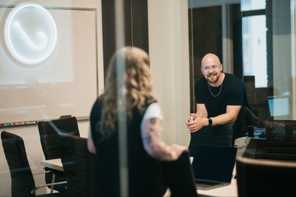 In a glass-walled conference room, two professionals discuss contractor marketing ROI; a laptop, whiteboard, and chairs are visible.