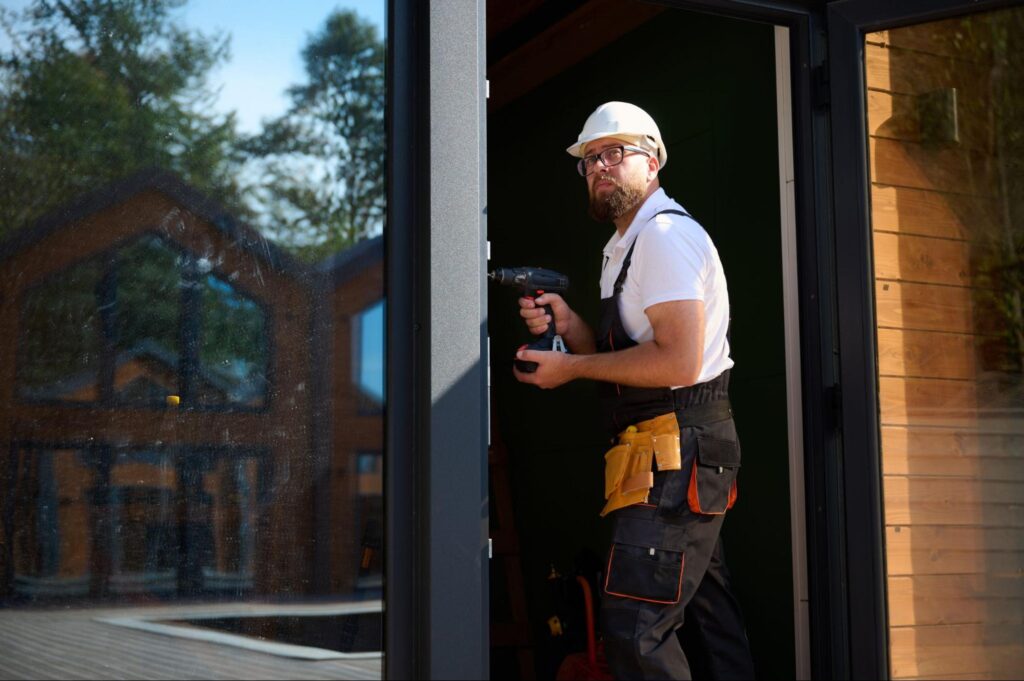 Construction worker in white hard hat and tool belt holds cordless drill in doorway; ideal image for window and door company marketing.