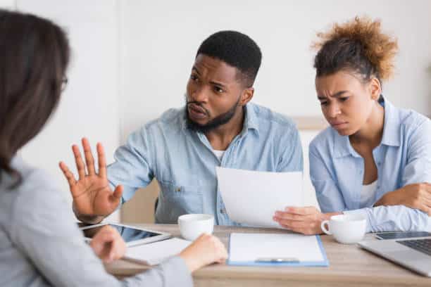 Two individuals sit at a table, reviewing roofing company documents with concern. The man raises his hand in a halting gesture as they discuss. A third person listens intently, while coffee cups and electronic devices are spread across the table.