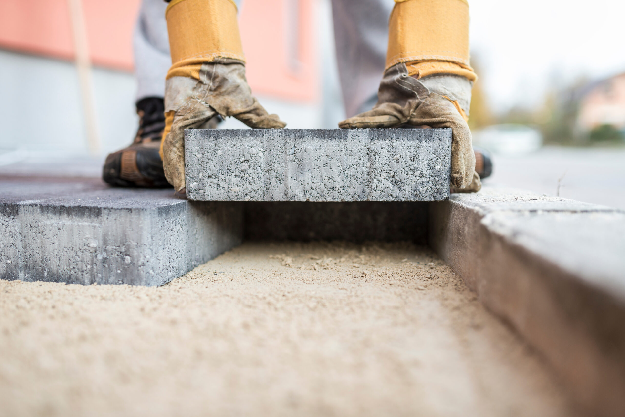 A skilled worker, clad in sturdy boots and protective gloves, expertly aligns a large concrete paving stone onto a sand base outdoors, ensuring precise placement among existing stones.