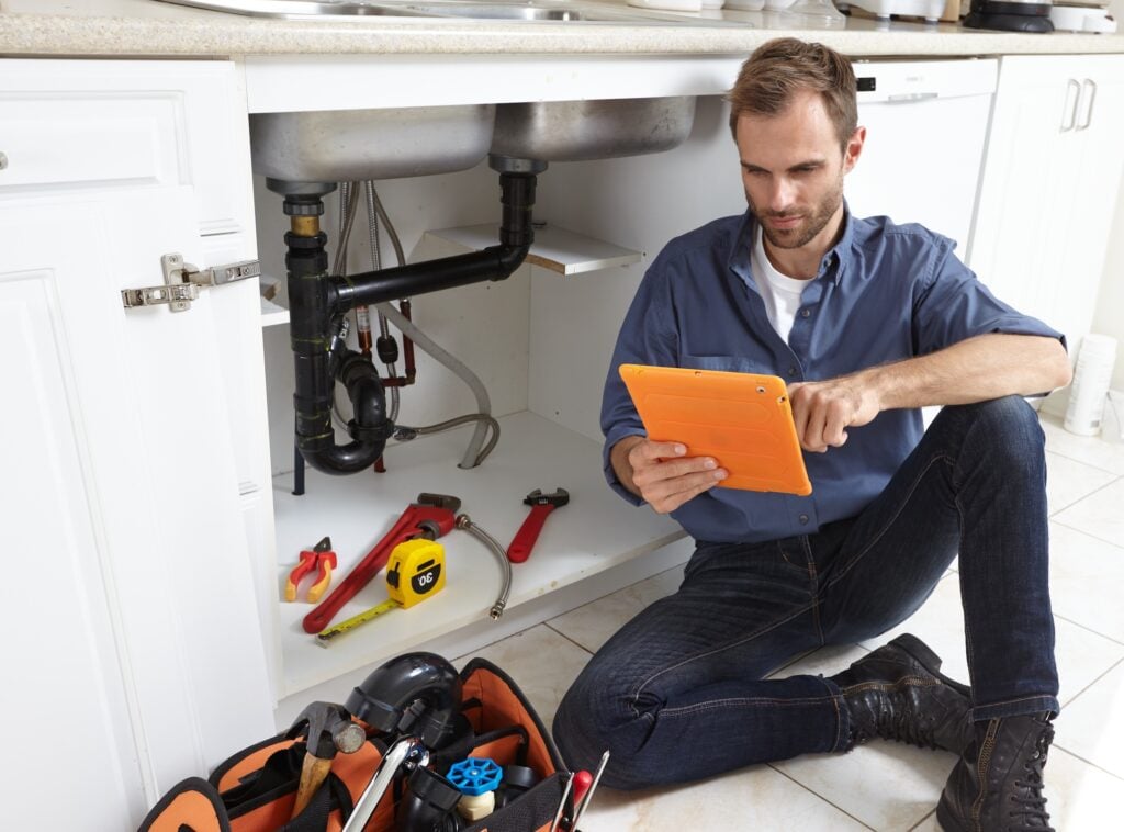 Plumber on kitchen floor with tools and tablet, researching top CRMs for 2025 to streamline repair job management—Hook Agency.