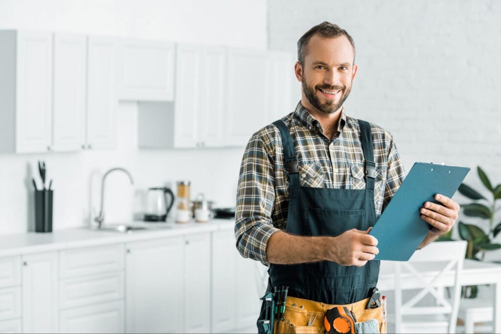 Handyman in plaid shirt and overalls with blue clipboard stands in bright kitchen, ready for plumbing work. Tools, sink, cabinets visible.