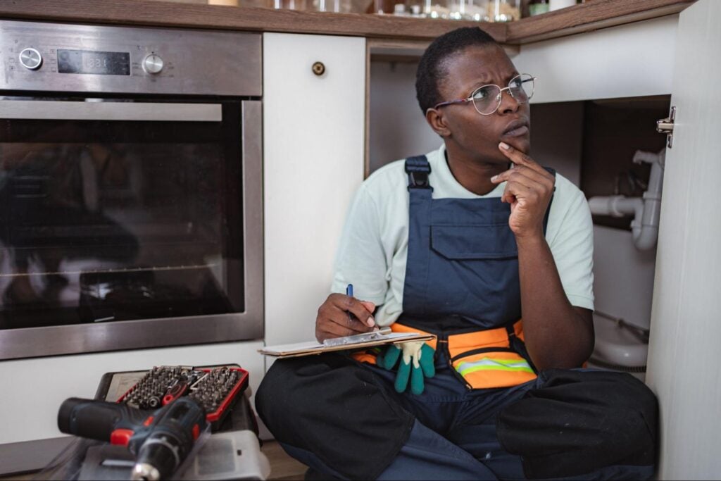 Plumbing marketing consultant in overalls and glasses reviews notes on kitchen floor, surrounded by tools and a drill.