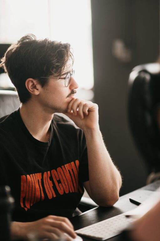 A focused individual in glasses and a mustache sits at their desk, hand thoughtfully on chin. Wearing a black "POINT OF CONTACT" T-shirt in bold red, they’re actively developing plumbing PPC strategies at the keyboard.