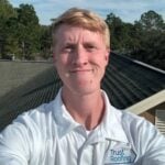 A Trust Roofing representative captures a selfie on a rooftop, showcasing the company's business growth. The image features them in a white polo shirt with the company logo, set against a backdrop of trees and a green-shingled roof under clear skies.