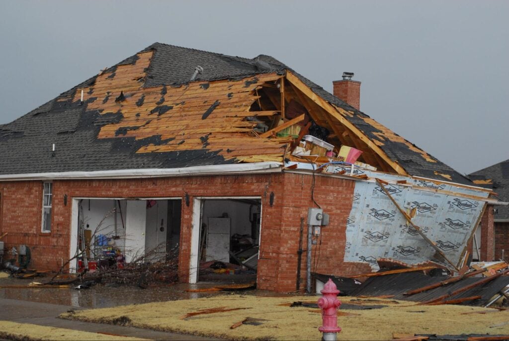 Brick house with major roof damage; missing shingles expose interior. Contractors repair as debris fills driveway. Pink hydrant visible.
