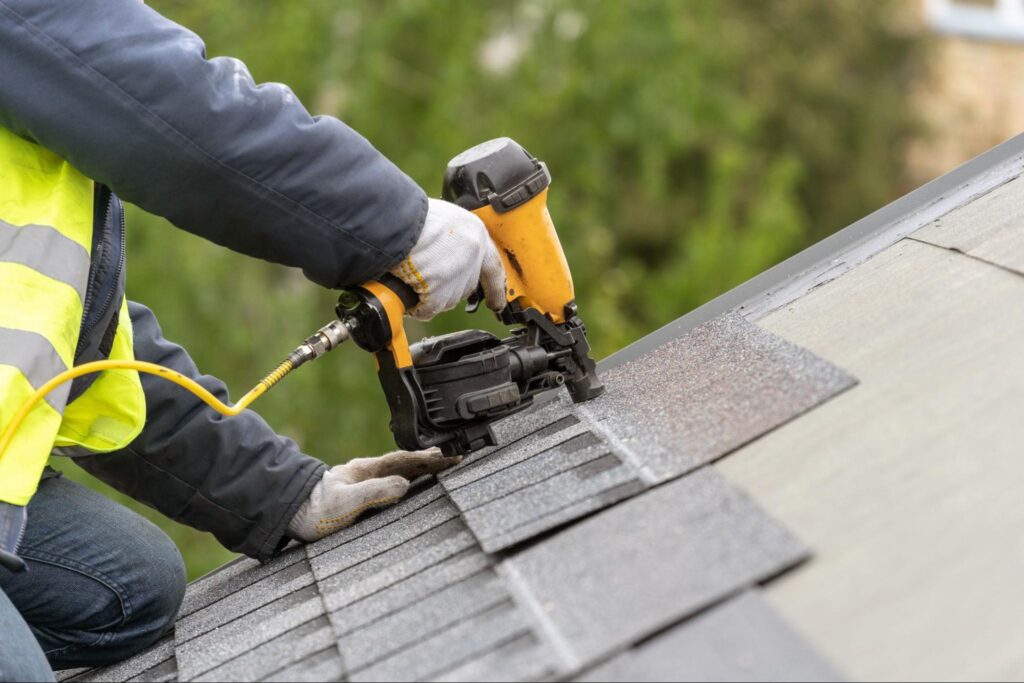 Homeowner in gloves and hi-vis jacket uses a nail gun to secure asphalt shingles on a sloped roof; green foliage visible behind.
