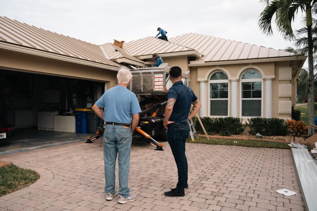 Two men observe as a roofing crew repairs the roof of a tan home—a familiar scene in high-demand home services industries. One crew member works atop the roof, while another operates from a lift near the garage. A palm tree frames the right side of the setting.