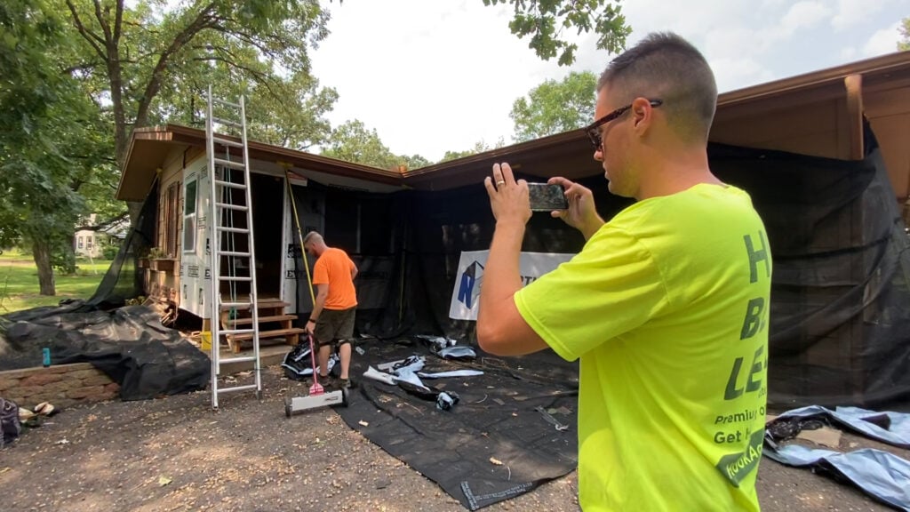 Man in yellow shirt records roofing renovation; ladders, tools, and materials highlight storm drought business success strategies.