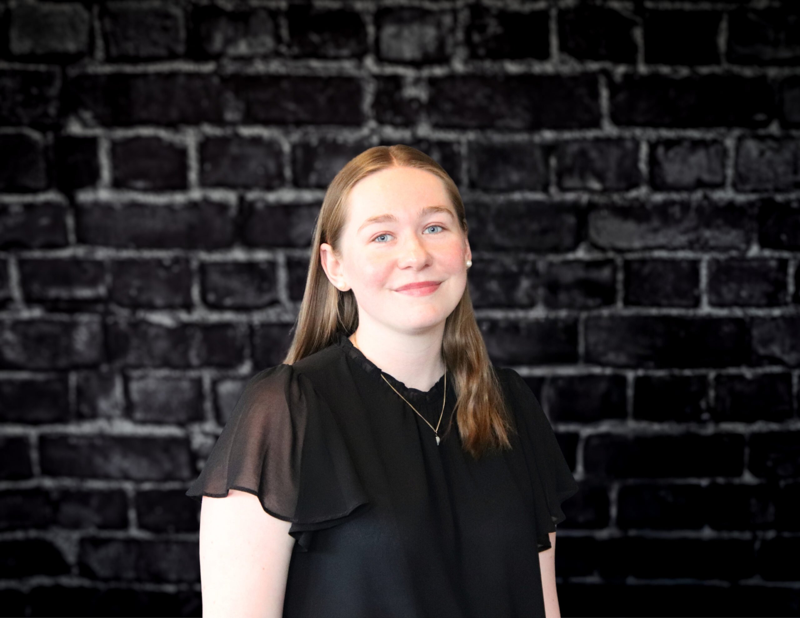 Selma Sandsengen, with long light brown hair, smiles in a black blouse and necklace before a dark brick wall on Hook Agency’s site.