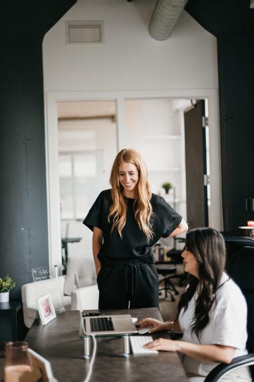 In a modern office setting, two women collaborate on developing solar marketing strategies. One stands confidently, dressed in black and smiling, while the other is seated at a desk with a laptop displaying their innovative approach. The office is elegantly designed with sleek decor and an inviting open door.
