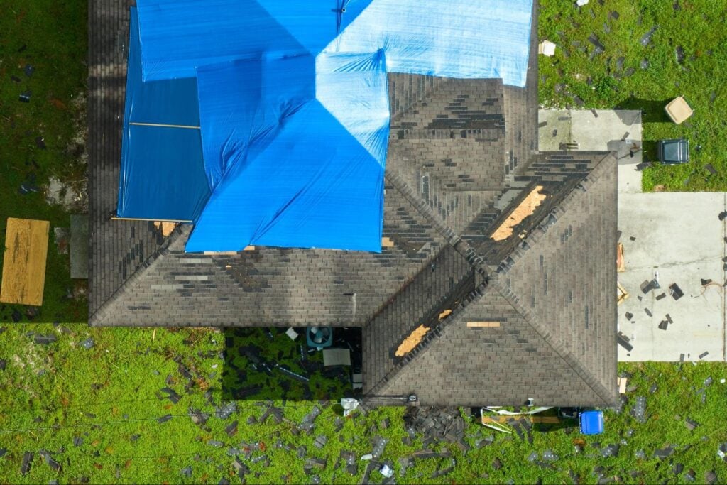 Aerial view of storm-damaged house with blue tarps on roof, debris and shingles on lawn—demonstrates urgent need for repairs.