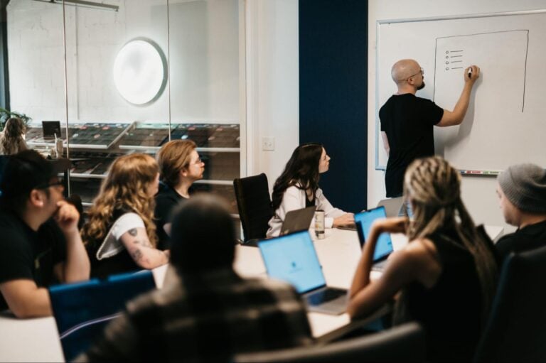 Team at conference table with laptops listens as a presenter outlines key steps on a whiteboard for vetting marketing agencies.