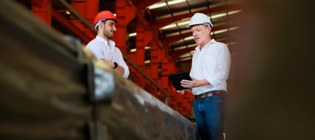 Two men in hard hats and white shirts stand inside a large industrial facility. One leads the discussion, referencing strategic management plans on a tablet, while the other listens attentively. Red beams and metal structures frame the background.