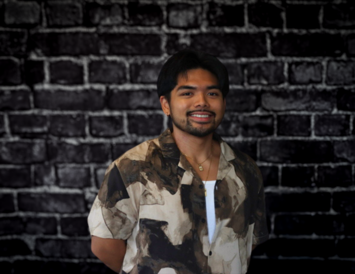 Axel Santo Domingo stands smiling before a black brick wall, wearing a patterned shirt, white undershirt, and gold necklace.