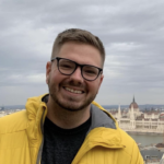 Chris Owens, wearing glasses and a yellow jacket, smiles confidently against a backdrop of a cityscape. The skyline showcases a historic building with a dome and spires beneath a cloudy sky.