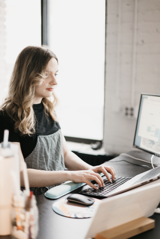 A woman with long hair, dressed in a black top and checked dress, diligently researches SEO strategies on her computer. Seated at a desk bathed in natural light from a large window, her focused gaze is fixed on the monitor.