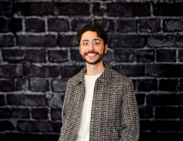 Smiling man with glasses, short dark hair, and trimmed beard in gray jacket and white shirt stands before black brick wall.