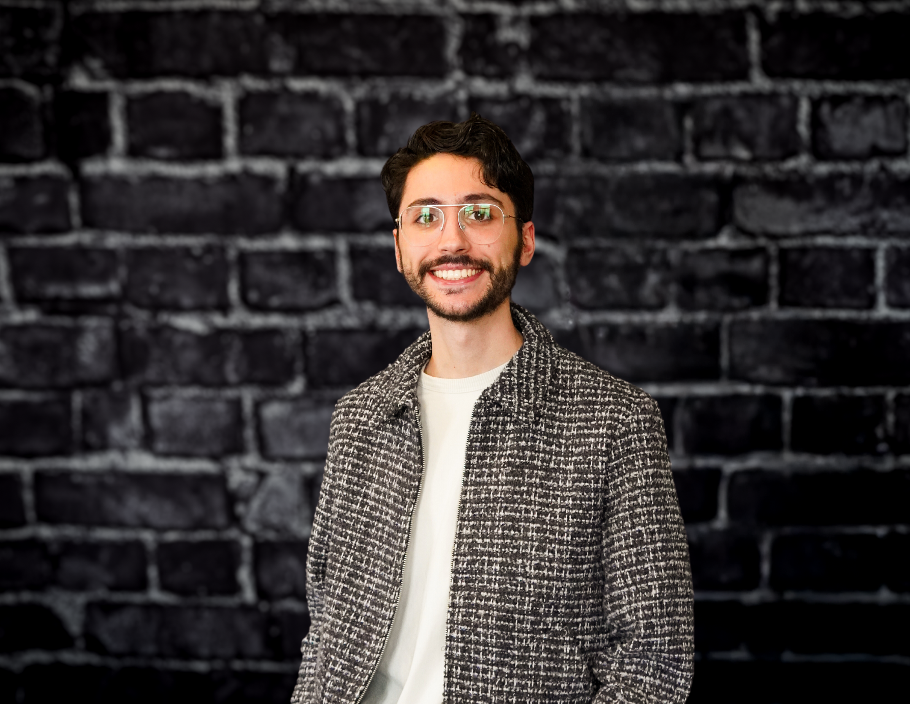 Smiling man with glasses, short dark hair, and trimmed beard in gray jacket and white shirt stands before black brick wall.