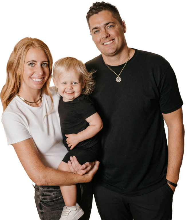 A smiling family stands against a plain background. The woman, with long blonde hair, holds a toddler dressed in black. Next to them, a man wearing a "Tree Service" T-shirt smiles warmly at the camera.