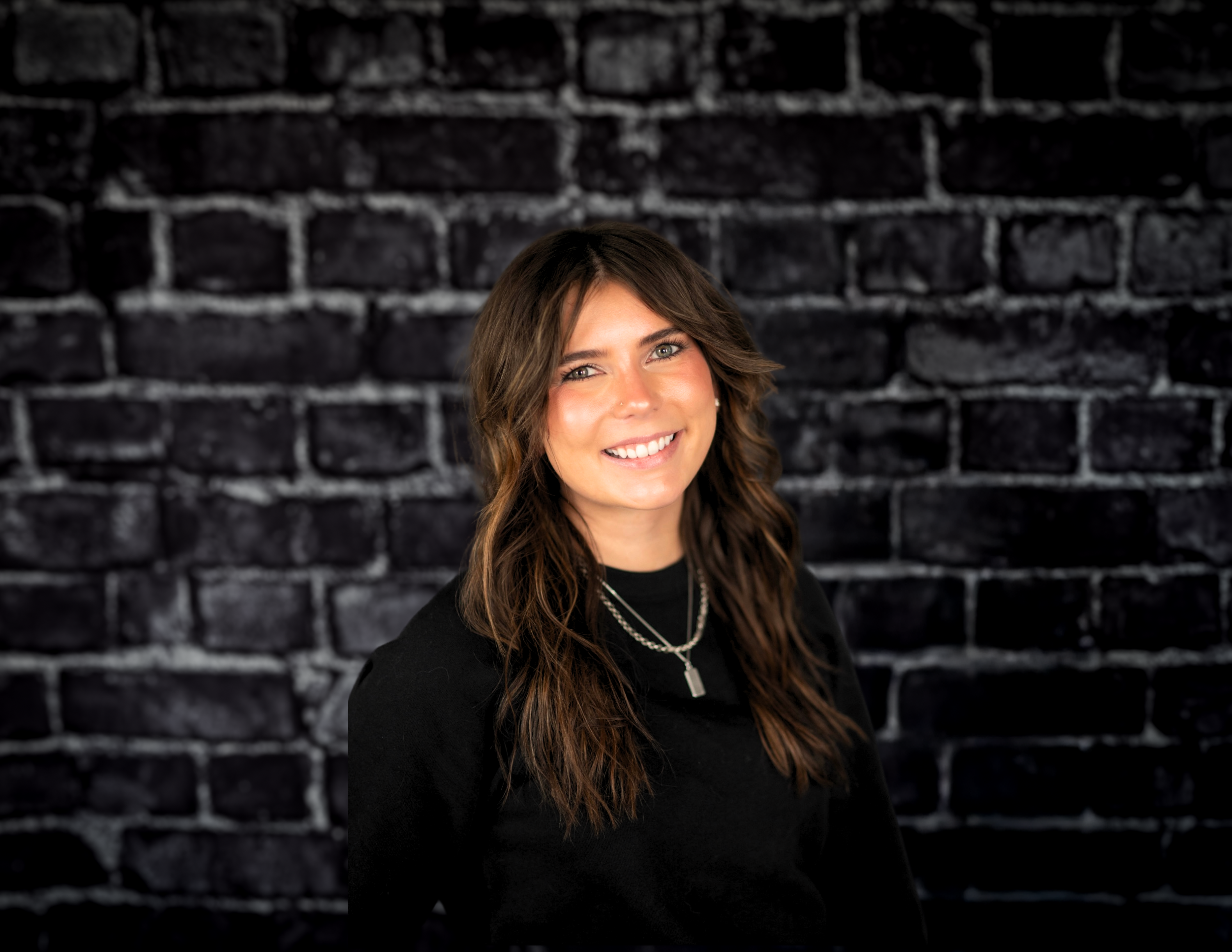 Brynn Wilson stands confidently against a dark brick wall, smiling warmly. She is wearing a black shirt complemented by two necklaces, her long brown hair cascading gently.