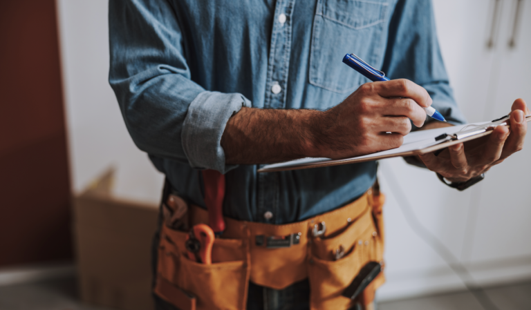 On Hook Agency's website, a professional in a denim shirt and tool belt embodies the essence of home services marketing by meticulously writing on a clipboard. The tool belt, filled with various tools, signifies preparedness for any challenge. A cardboard box is subtly visible in the background.