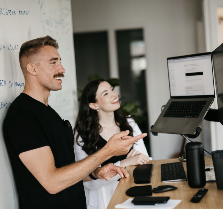 Two individuals stand at a desk, strategizing Home Services SEO with focused expressions. A whiteboard filled with notes serves as their backdrop, while the desk is equipped with tech gadgets including a tablet and smartphone, fostering an innovative workspace.