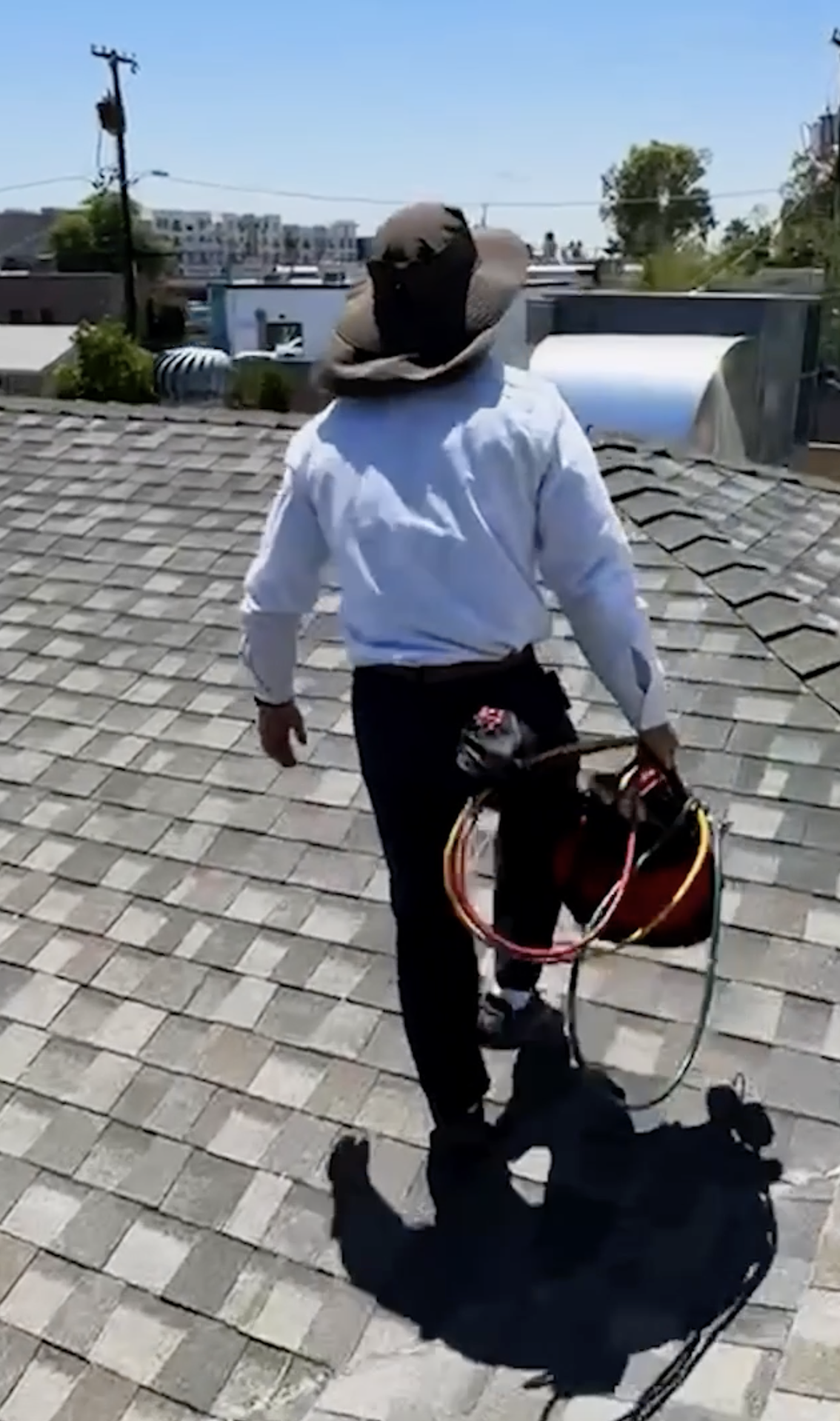 A professional in a wide-brim hat, white shirt, and dark pants carries a tool bag and coiled cables while walking on a shingled roof—an ideal visual for home services marketing. Buildings and trees with a clear sky complete the background.