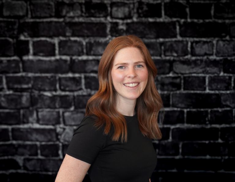 Megan Talbot, Hook Agency team member with long wavy red hair and a black t-shirt, smiles before a dark brick wall.