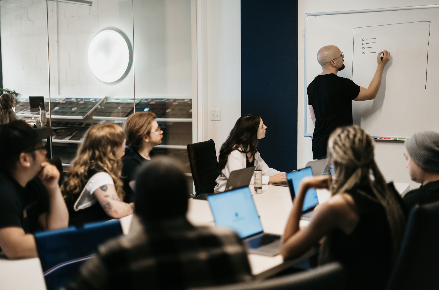 In a glass-walled meeting room, a focused team observes as their leader outlines SEO strategies on the whiteboard, fostering an atmosphere charged with concentration and innovation.