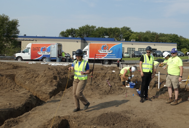 At a bustling construction site, workers clad in reflective vests and hard hats engage in focused tasks. A plumbing company team member strides by, while colleagues efficiently manage trench work. Nearby, two branded trucks are stationed amidst the serene backdrop of a building and trees under a clear sky. Plumbing marketing company Hook agency out in the field helping to understand their clients so they can market better.