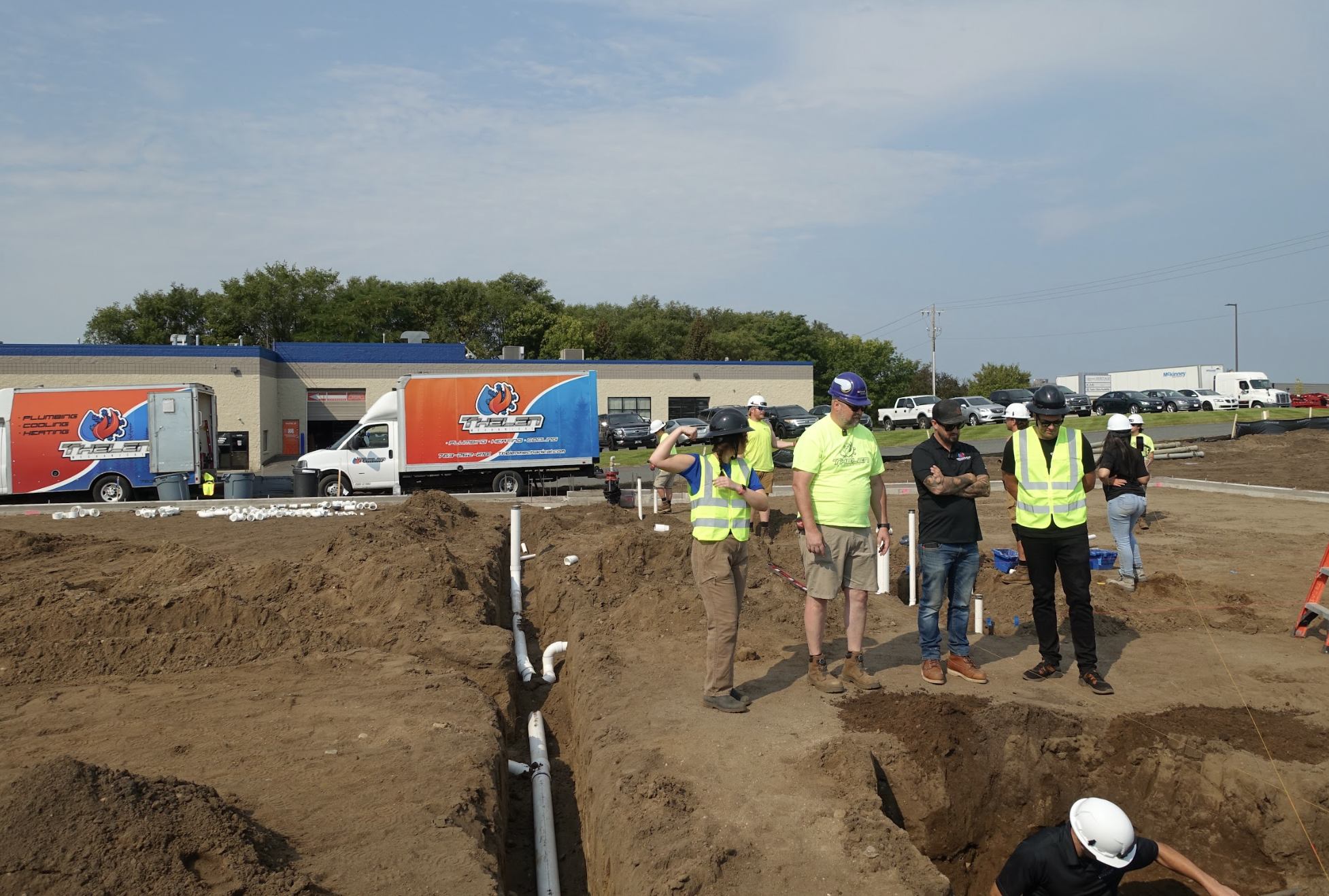On a busy construction site, skilled workers in safety gear operate near a trench installing advanced plumbing systems. Nearby, trucks with vibrant logos are positioned as a new building emerges against the cloudy sky.