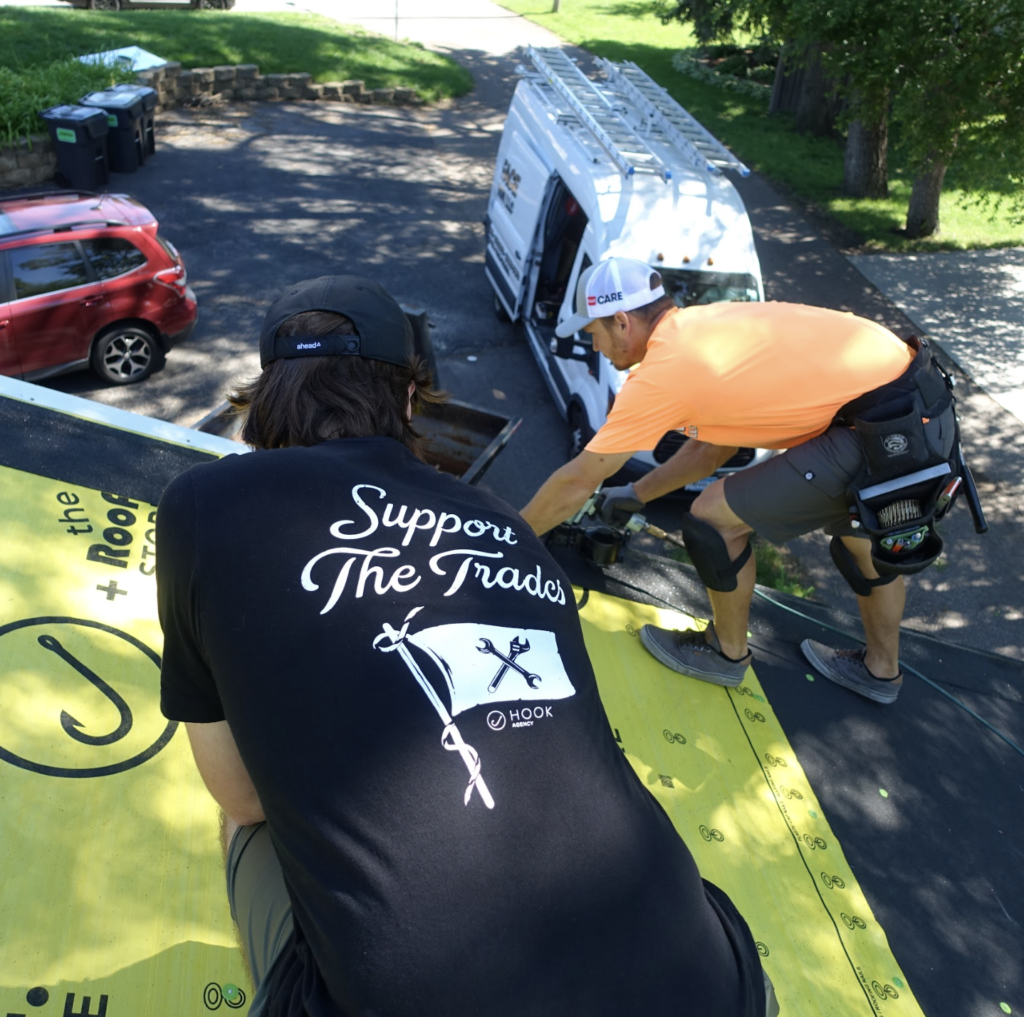 Two workers install roofing material, ideal for a home services video. In the foreground, one wears a black "Support the Trades" shirt. Nearby, another in orange kneels with a tool belt at the roof's edge. Below, a white van and red car stand ready for their next task - roofing and home services video marketing tips and strategies that actually work.