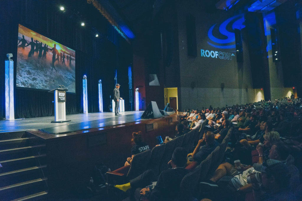 Speaker at ROOFCON addresses growth and company culture; event logo and beach running image are displayed behind on stage.