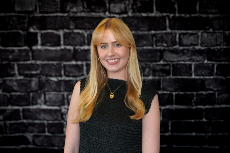 A professional woman with long blonde hair and a gold pendant necklace stands smiling confidently against a black brick wall.